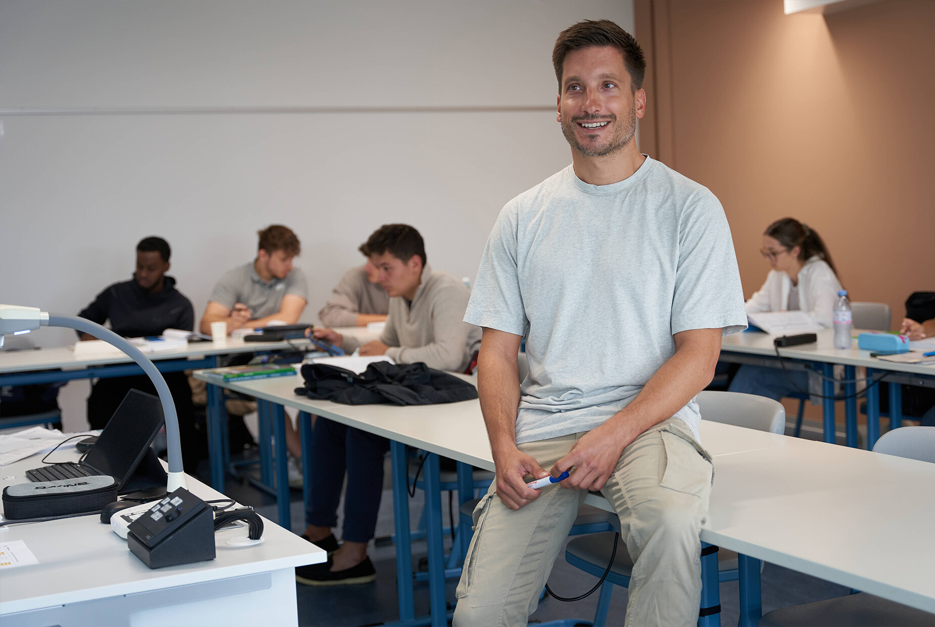 Im Vordergrund sitzt eine Lehrperson auf einem Pult. Im Hintergrund sitzen mehrere Schüler/-innen an Pulten.