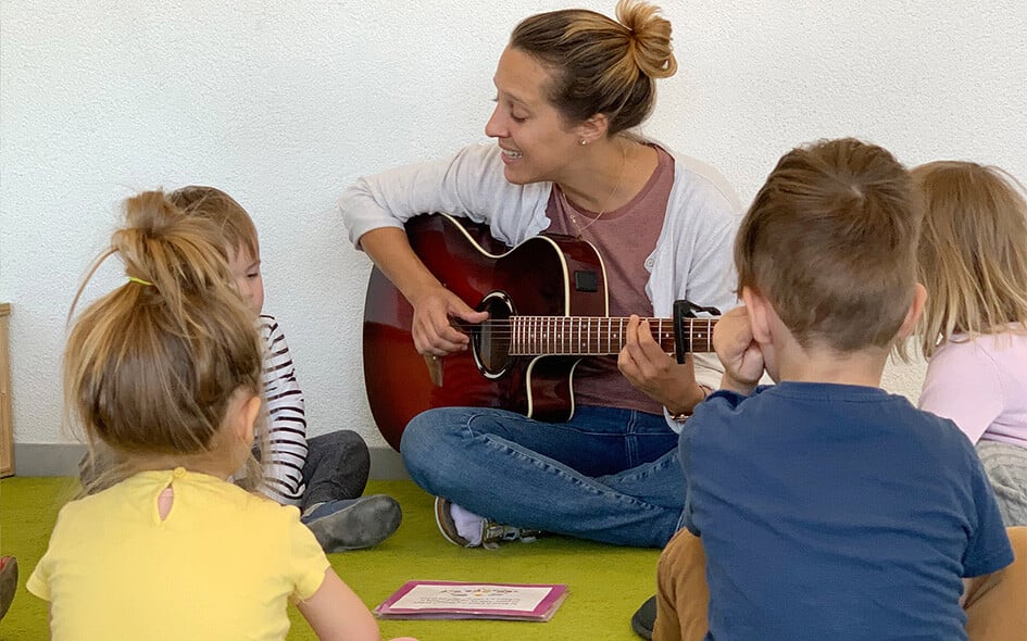 Eine Person mit Gitarre sitzt am Boden mit vier Kindern.
