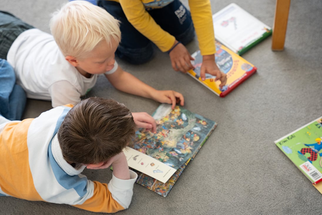 Drei Kinder liegen oder sitzen am Boden und schauen ein Bilderbuch an.