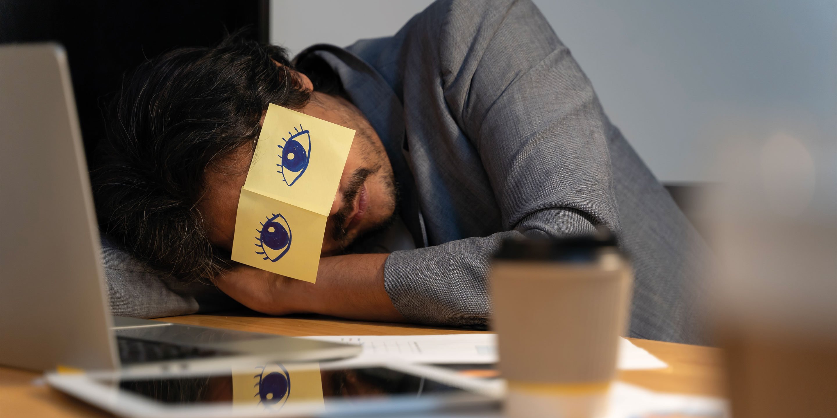 Eine Person liegt mit dem Kopf auf einem Tisch. Über den Augen eine Zeichnung mit einem Augenpaar.