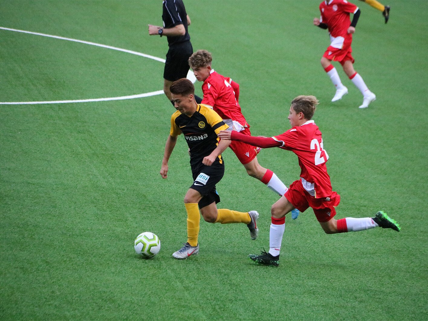 Ein Fussballspieler in einem gelb-schwarzen Trikot und zwei Fussballspieler in einem roten Trikot kämpfen um den Ball.