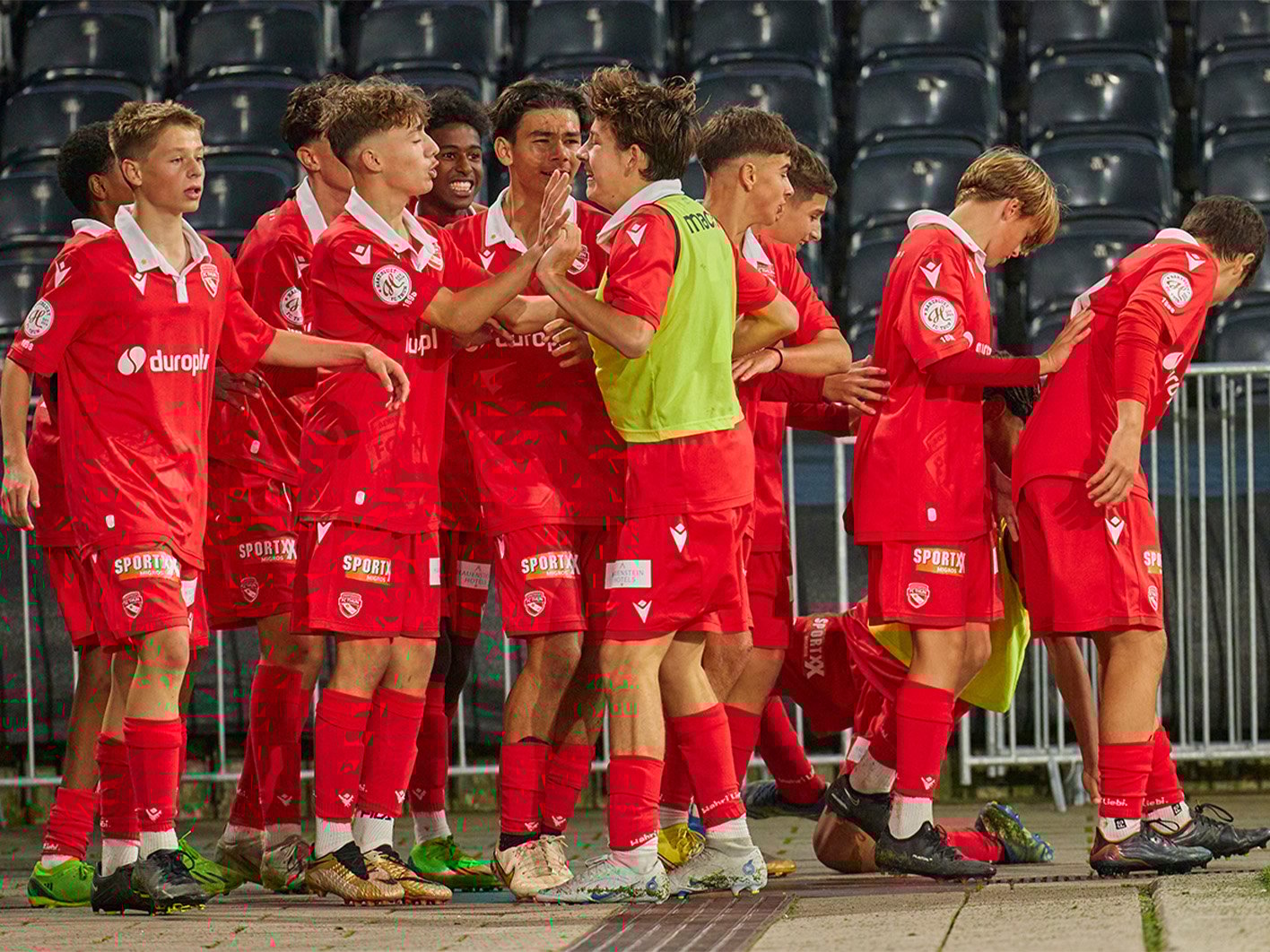 Verschiedene Spieler in roten Trikots stehen neben dem Fussballfeld.