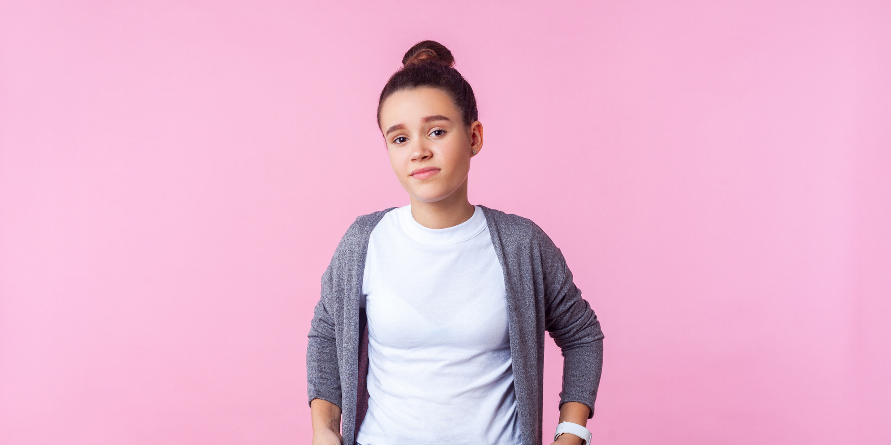 Eine junge Frau mit einem weissen T-Shirt und einer grauen Jacke steht vor einem rosa Hintergrundbild, die Hände in den Hosentaschen.