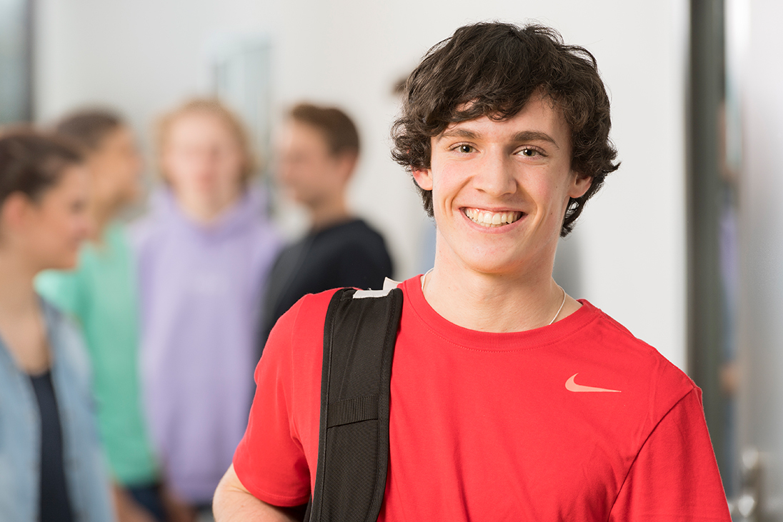 Lächelnder Schüler im roten Sportshirt mit Rucksack vor einer Gruppe Jugendlicher.