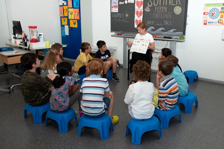 Die Kinder sitzen in einem Kreis. Die Lehrerin erzählt aus einem Arbeitsbuch.