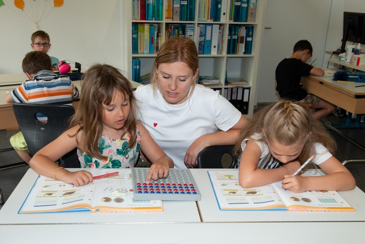 Im Vordergrund sitzen 2 Mädchen an einem Pult. Vor ihnen liegt das geöffnete Arbeitsbuch. Die Lehrerin erklärt eine Aufgabe.