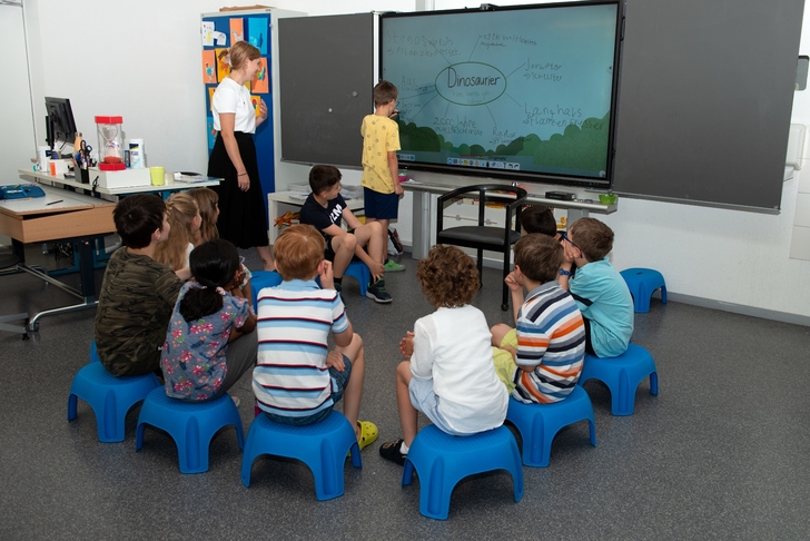 Die Kinder sitzen in einem Halbkreis. Ein Schüler schreibt an die interaktive Wandtafel. Die Lehrerin unterstützt den Schüler.