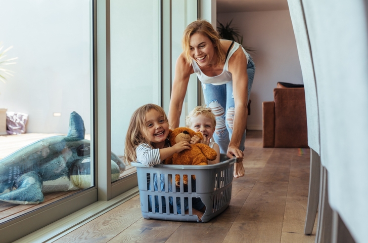 Eine Frau schiebt zwei Kinder in einem Wäschekorb am Boden entlang.