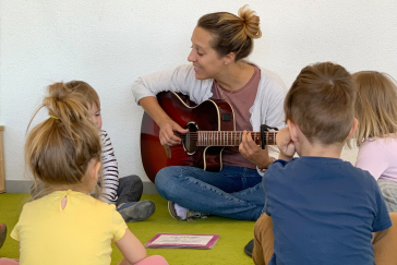 Frau spielt Gitarre. 4 Kinder hören zu.
