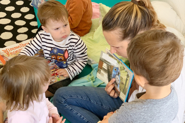 A woman is sitting on the floor showing a picture book to three children..