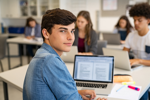 Ein Junge sitzt an einem Pult und schaut schräg über die Schulter in die Kamera. Er arbeitet mit einem Laptop.